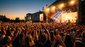 Tłum z rękami w górze na festiwalu przy Galerii Jurajskiej w Częstochowie. Rozświetlona scena i jupitery o zachodzie słońca podczas letniej imprezy plenerowej.